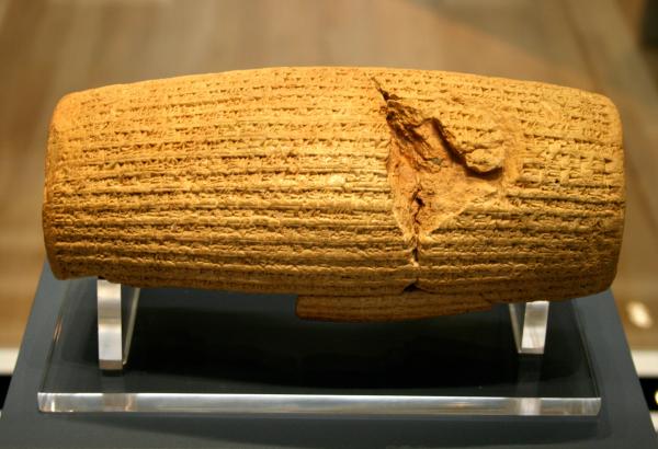 A cylindrical scroll with grooves laid to the side mounted on a museum display; there is a large crack on the right side of the scroll.