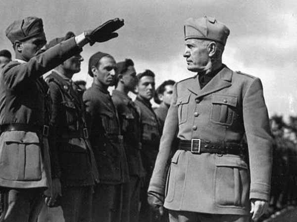 Black and white photo of Mussolini inspecting a line of soldiers while wearing uniform and hat. A soldier extends his right arm and gloved hand in salute